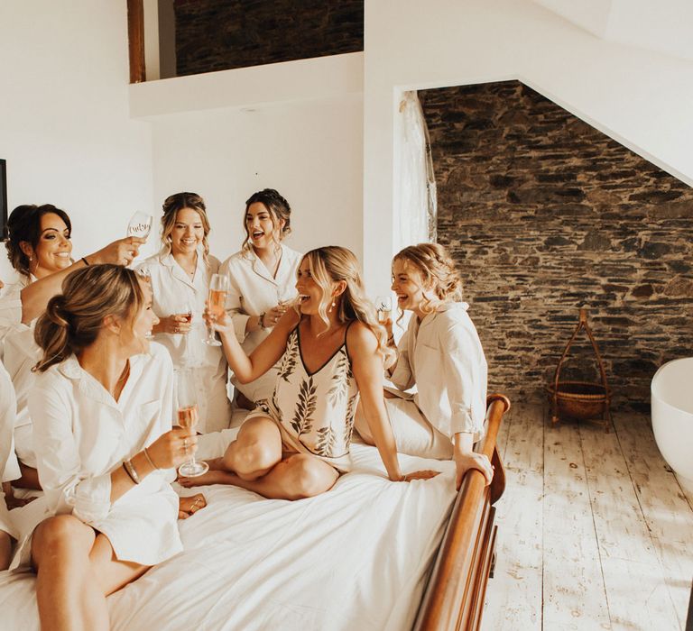 Bride in cami pyjama set and bridesmaids all in long white shirts raise flutes of sparkling wine as they sit on large bed before wedding at Anran Devon