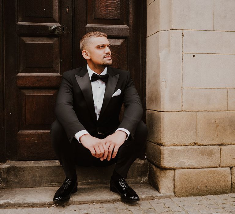 Stylish groom sitting on a step in a Reiss tuxedo with satin lapels, bow tie and shiny shoes 