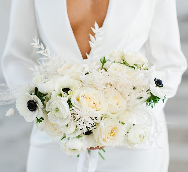 A bride wears a low cut white suit jacket and holds a bouquet filled with white flowers and anemones.