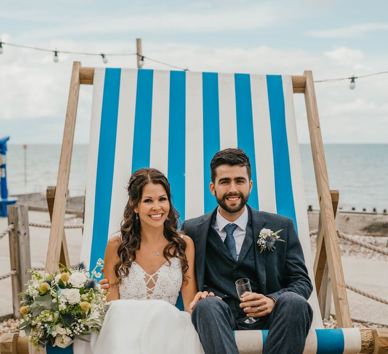 Bride & groom sit on giant foldable chair in blue and white on the beach front on their wedding day