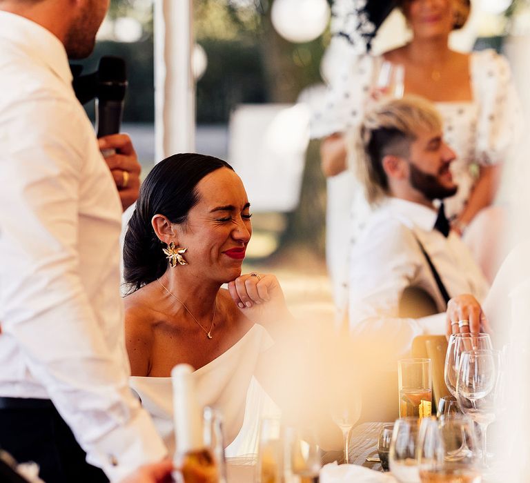 Bride in gold flower earrings laughs during wedding speeches