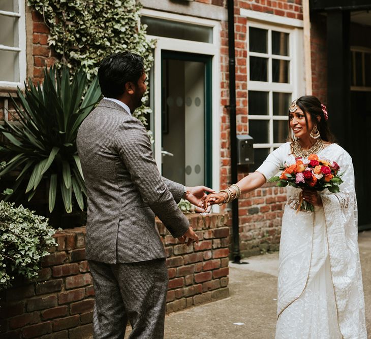 A first look for Indian British wedding. The groom wears a grey suit and the bride wears a white sari and carries a colourful bouquet.