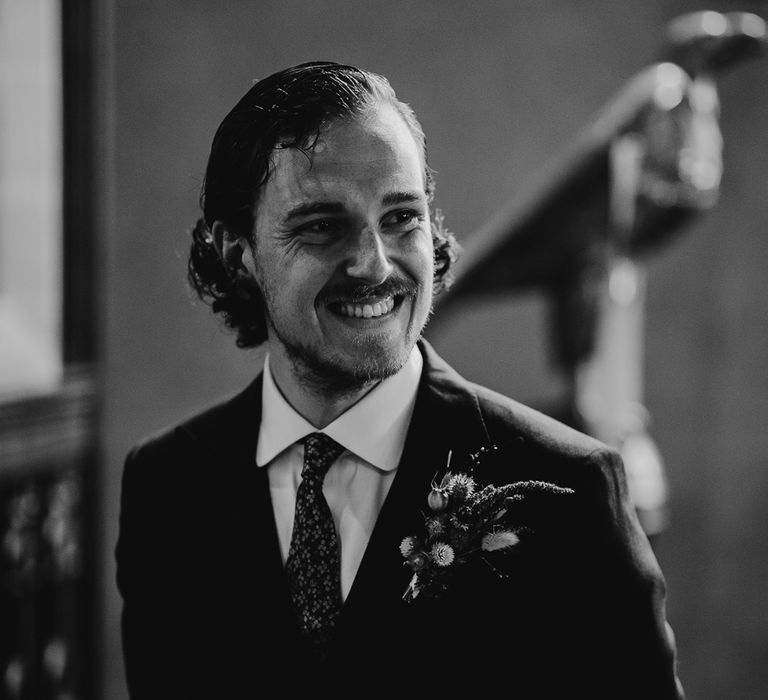 Groom in three piece suit, patterned tie and dried floral buttonhole smiles as he waits for brides during church wedding ceremony in Cornwall