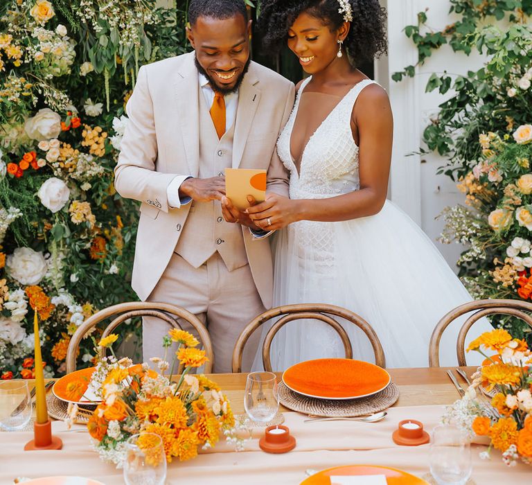 Bride in a plunging neckline wedding dress reading the wedding menu with her groom at their orange decorated outdoor reception 