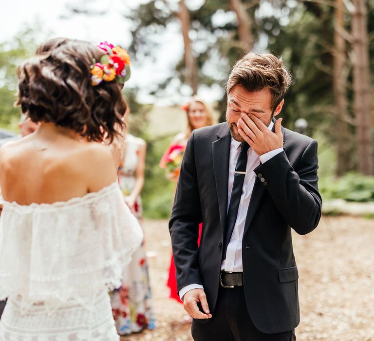 Groom in black suit wipes his eyes during outdoor wedding ceremony with bride in off the shoulder Grace Loves Lace wedding dress and colourful flower crown