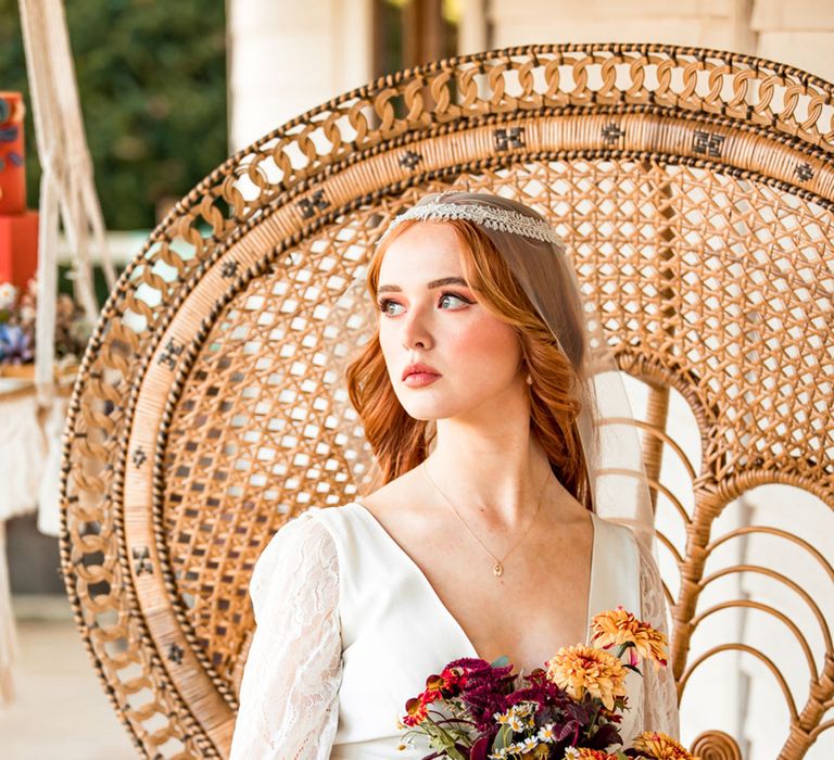 Bride in long lace sleeve wedding dress and Juliet cap wedding veil holding colourful bridal bouquet sits in large rattan chair on veranda