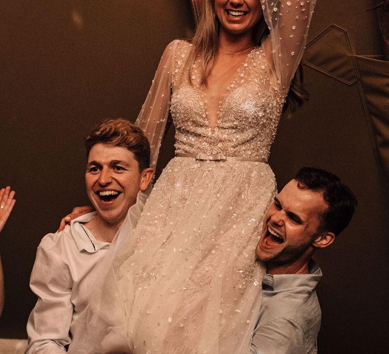 Bride in long mesh sleeve embroidered wedding dress and flower crown is held up on shoulders of two wedding guests at tipi wedding reception