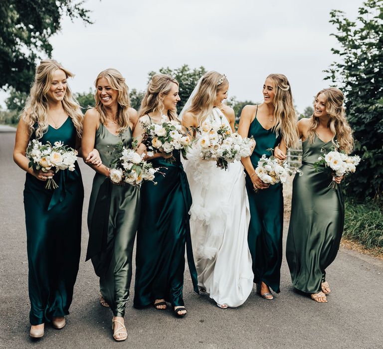 Bride in Halfpenny London wedding dress and veil walks down road with bridesmaid in green satin bridesmaid dresses all holding white and green wedding bouquets 