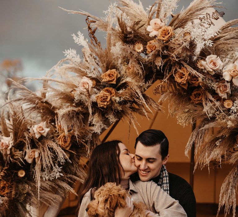 Bride and groom-to-be embracing in their bell tent decorated with dried flowers with their pet cockapoo