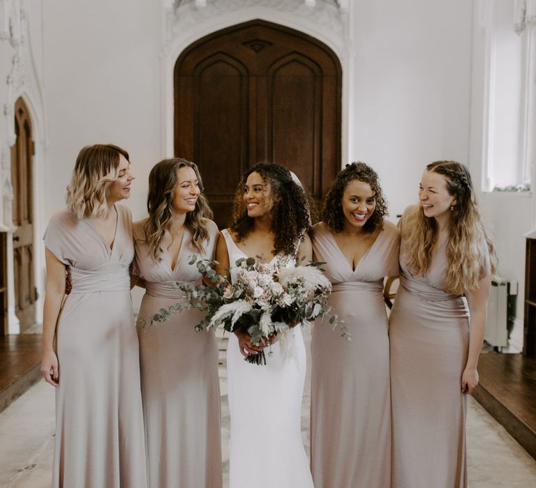 Bride stands with her bridesmaids who wear neutral bridesmaid gowns with criss-crossed waistbands