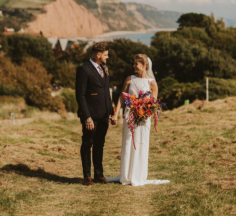 Coastal wedding with bride in Charlie Brear wedding dress and cathedral length veil holding a bright pink wedding bouquet and her grooms hand in a navy suit 