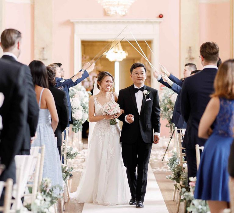 Bride walks down the aisle with her father who wears black tie as swords are held above them for Military ceremony