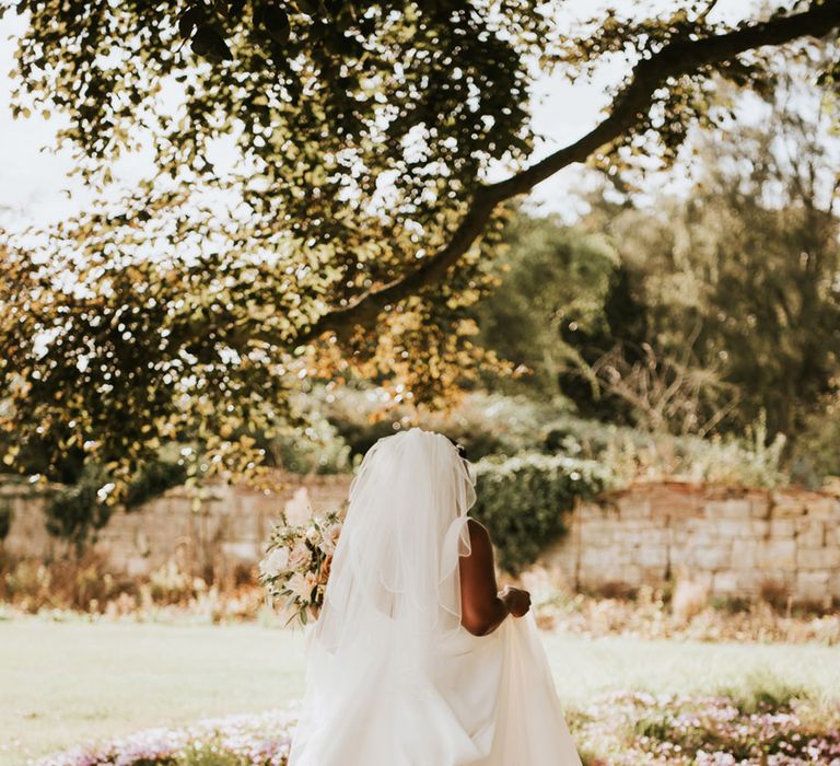 Black bride in an Essense of Australia wedding dress and cathedral length veil walking through the flower bed 