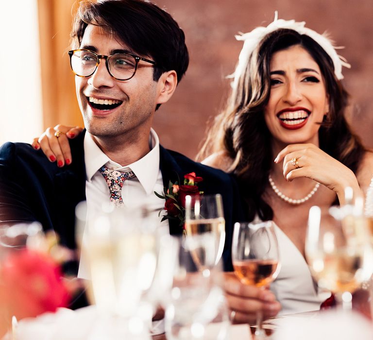 Laughing bride in pearl necklace and feathered headband sits with groom in dark suit and floral tie during Elmore Court wedding breakfast