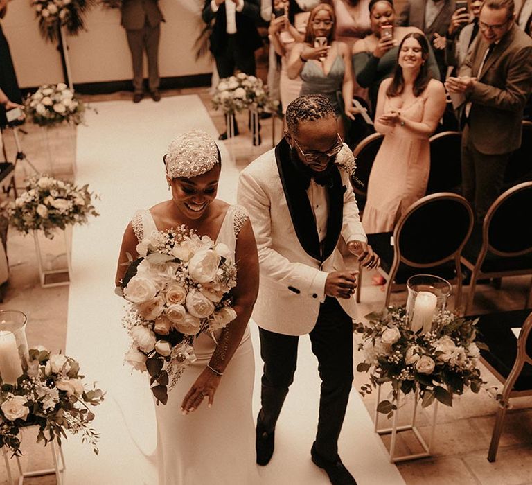 Intimate wedding ceremony with Black bride in a vintage style wedding dress and beaded headdress descending up the aisle with her groom in a white tuxedo jacket 