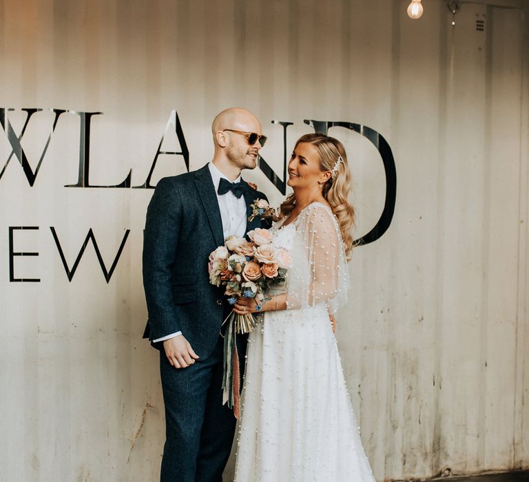 Bride in pearl wedding dress with sleeves and train holding pastel mixed rose wedding bouquet stands with groom in blue tweed suit with bow tie