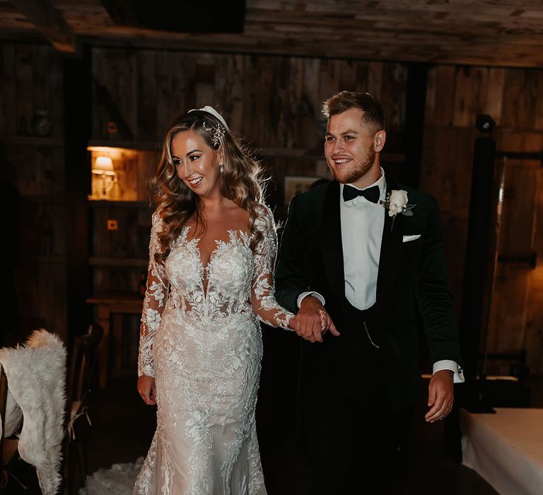 Bride in illusion lace fitted wedding dress walking hand in hand with groom in black tie green velvet suit jacket 