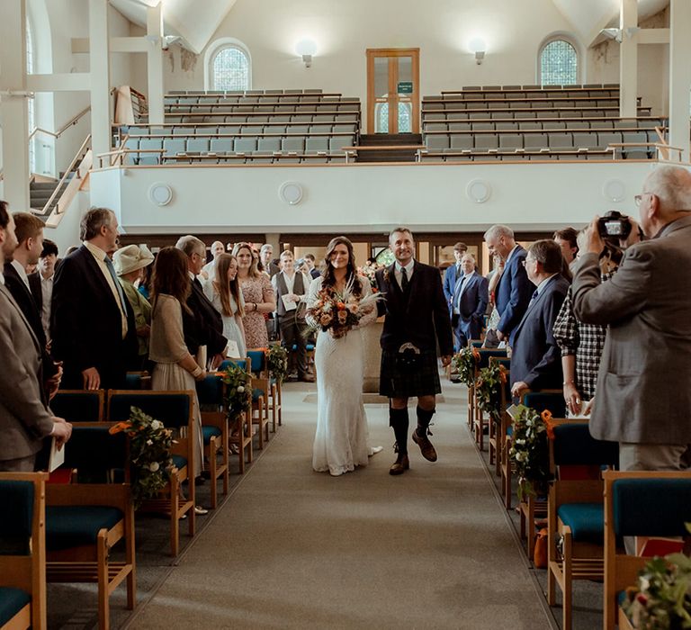 Father of the bride in traditional kilt walks the bride down the aisle as all guests stand and take pictures