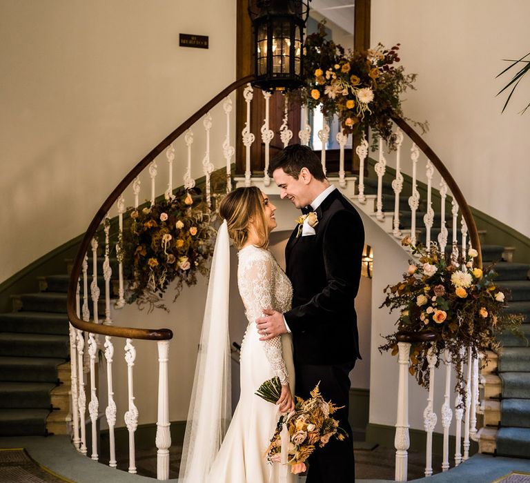 Bride in lace top long sleeve fitted wedding dress with train and buttons with groom in black tie with autumnal flowers 