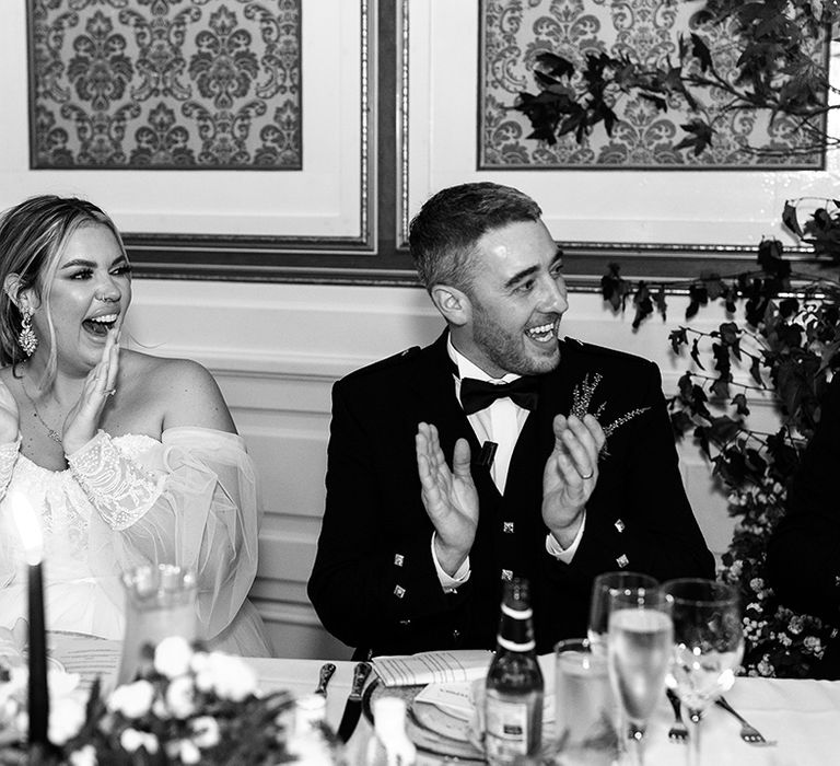 Bride in strapless wedding dress with sheer sleeves and lace cuffs and groom in black tie applaud for speeches