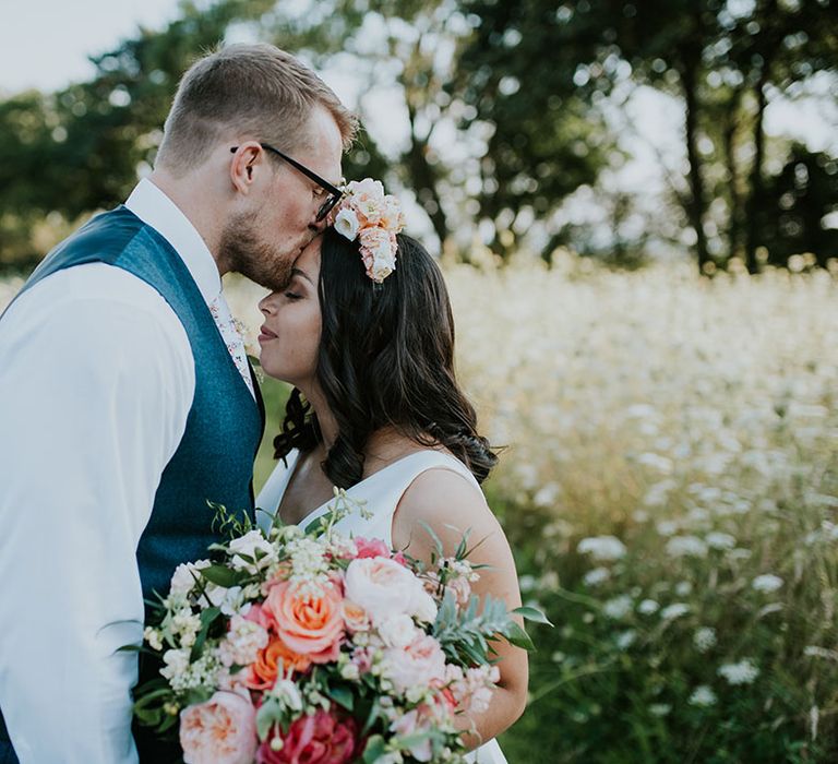 Groom in blue waistcoat kisses the brides forehead who carries a bright wedding bouquet for summer wedding