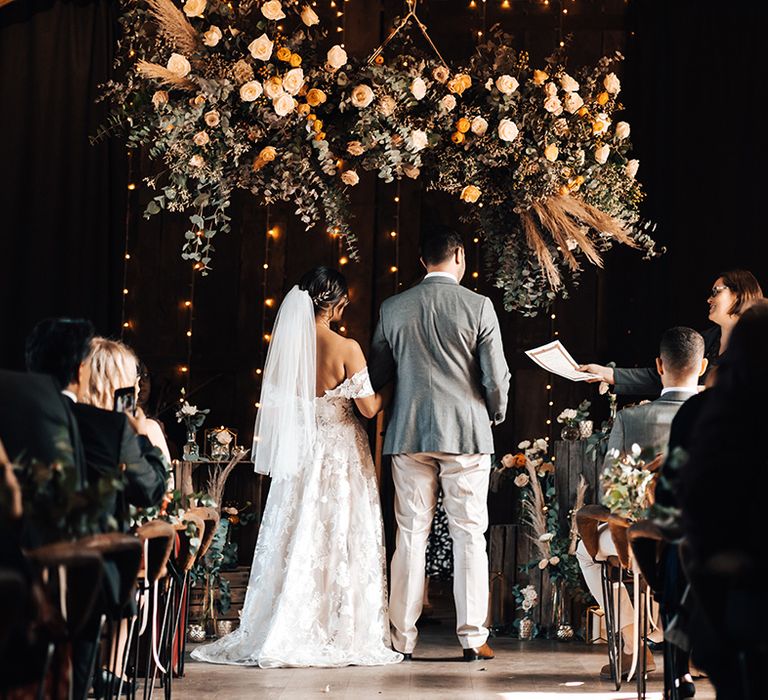 Bride in floral lace dress and groom in grey suit stand at the altar of their civil ceremony at Tythe Barn