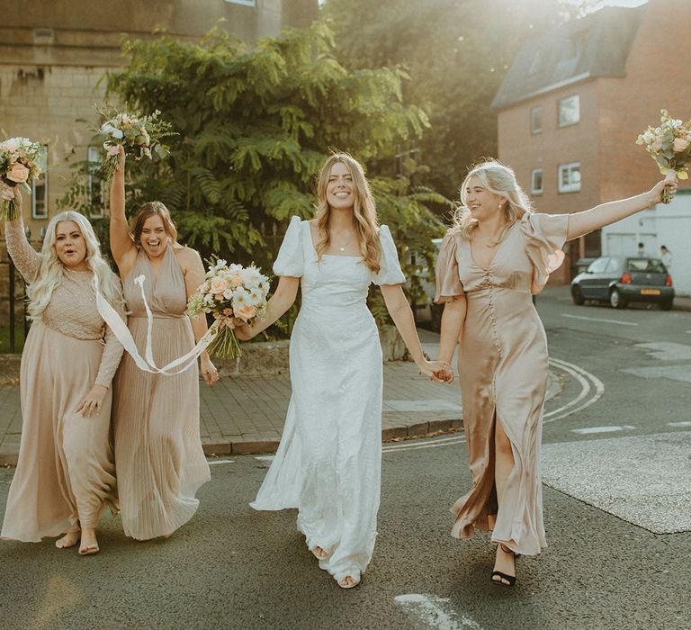 Bride in Story of My Dress gown holding pink and white bouquet walking with bridesmaids in different style champagne gold dresses 