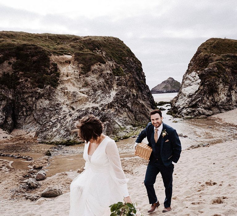 Bride walks barefoot in the sand in her Sassi Holford wedding dress as groom walks behind in blue suit carrying wicker picnic basket 