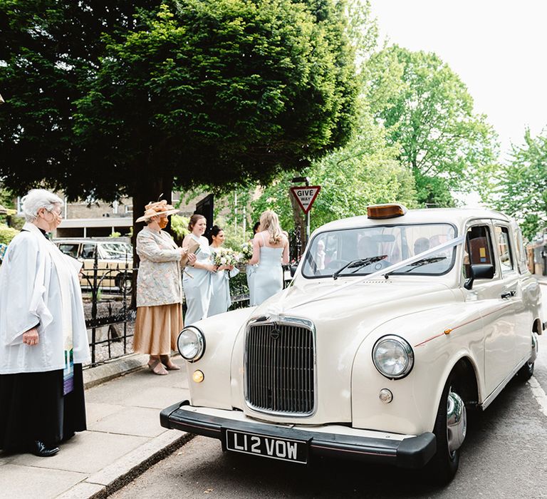 Wedding guests and bridesmaids wait as a cream taxi cab decorated with ribbons pulls up with the bride