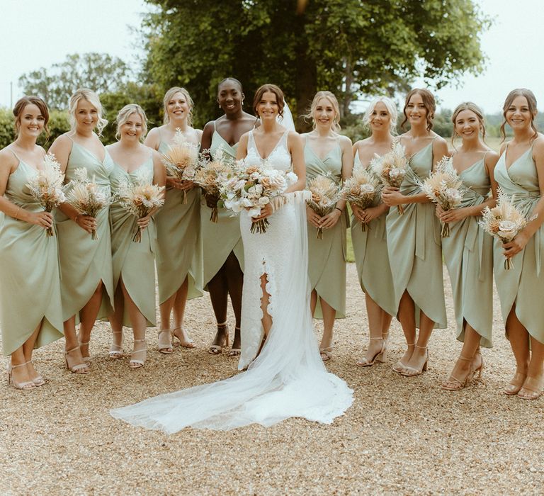 Bride in v-neck lace wedding dress with front slit stands with her bridesmaids in sage green wrap dresses and dried flower bouquets