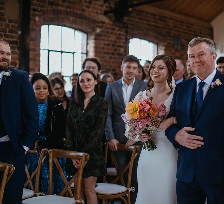 Bride holding bright colourful spring wedding bouquet with giant poppy is walked down the aisle by father of the bride in navy blue suit and tie