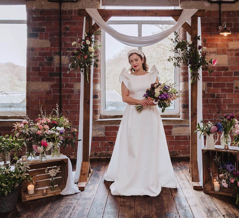 Bride in Jesus Peiro wedding dress with raised sleeves smiling at the altar with wooden frame and wedding drapery surrounded by colourful flowers for spring wedding