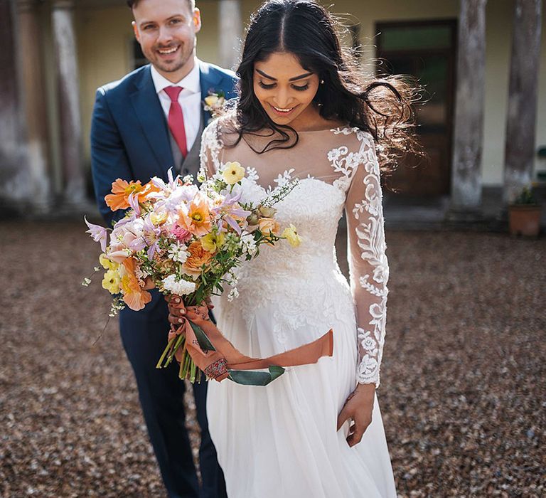 Bride in sweetheart illusion lace wedding dress holding spring wedding bouquet is followed by groom in navy blue suit and red tie