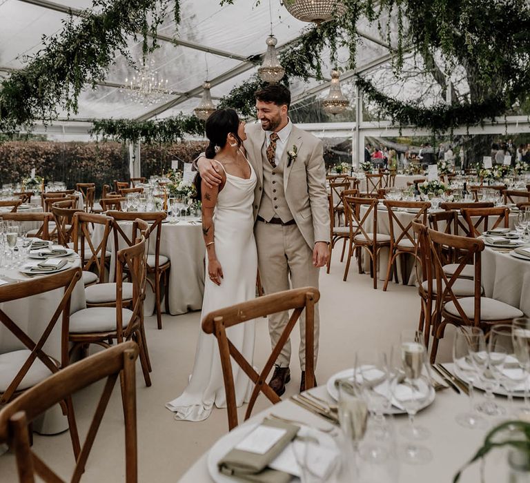 Bride in a satin wedding dress and groom in a beige suit standing in the middle of their clear glass marquee wedding reception with wooden furniture, greenery decor and handing chandelier 