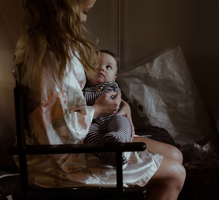 Bride in satin floral robe sits with baby in striped baby grow 