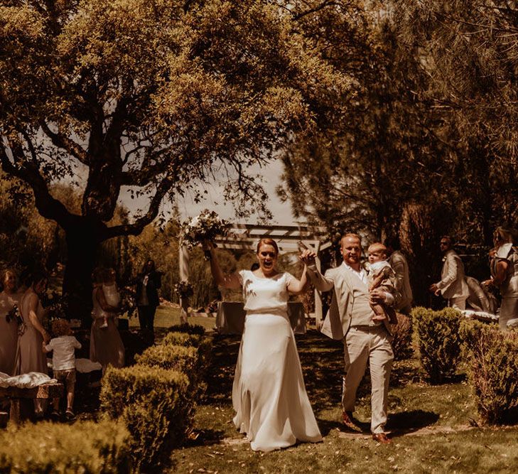 Bride & groom walk along grass and carry their baby after outdoor wedding ceremony in Spain 