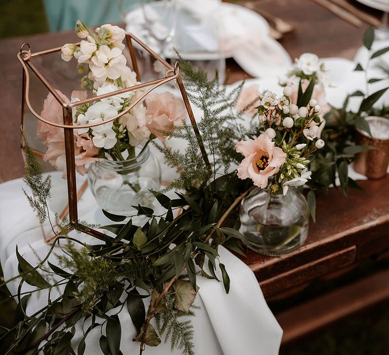 Green foliage table runner, white fabric and gold glass lanterns filled with pink flower stems wedding table decor 