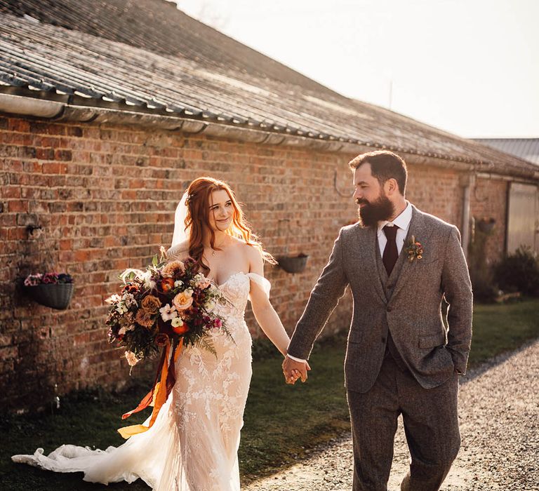 Bride in off the shoulder fitted wedding dress carrying an autumnal coloured bouquet with groom in a grey suit and burgundy tie 