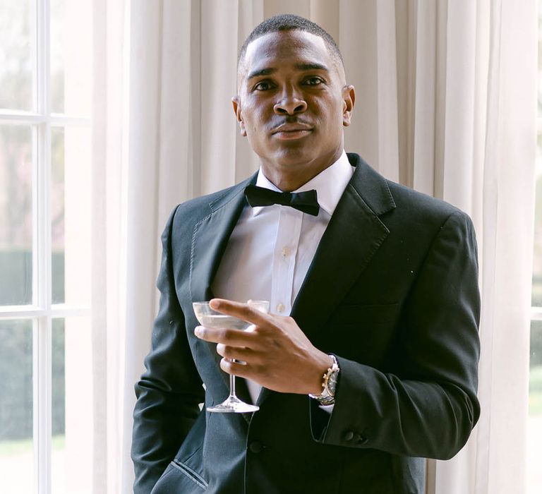 Groom stands looking at the camera in black tie with a cocktail glass 