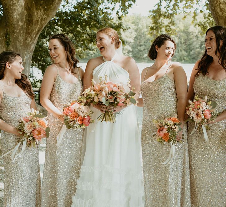 Bride wearing London Halfpenny wedding dress and carrying pale pink peony floral bouquet stands with her bridesmaid in gold sparkly bridesmaid dresses