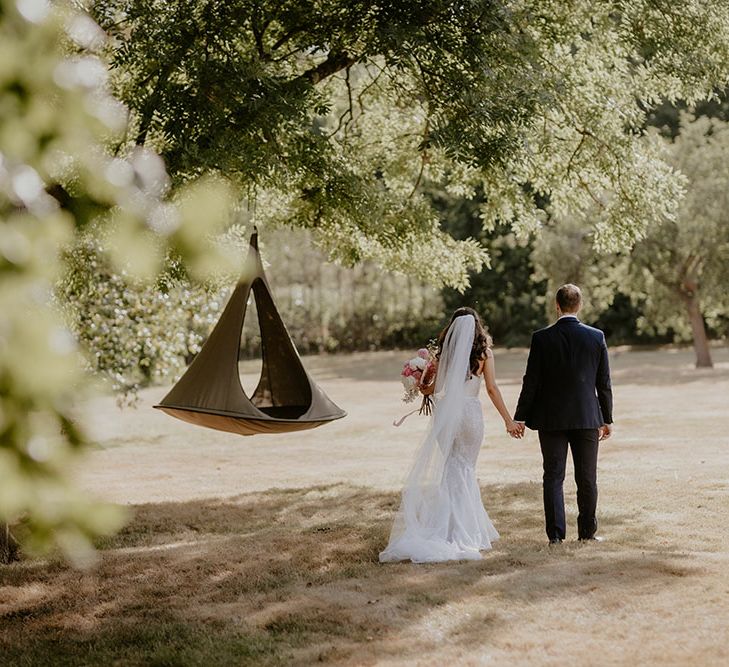 Bride wears floor-length veil and walks with her groom in family garden 