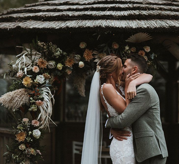 Bride & groom have their first kiss during wedding ceremony as champagne and rust florals line the front of the beach hut 