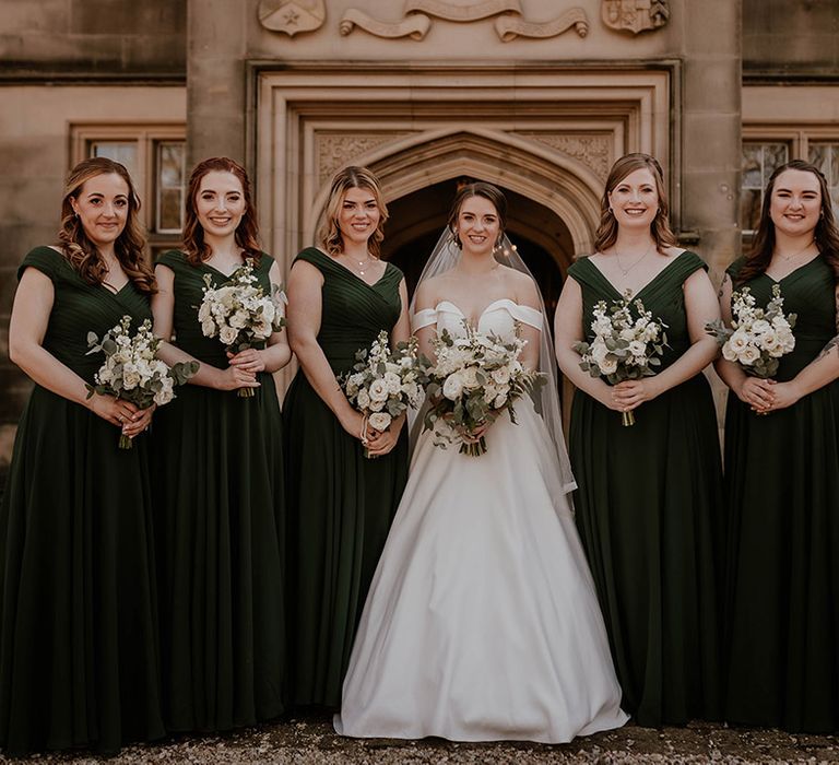 Bridesmaids in dark forest green dresses with the bride in an off the shoulder wedding dress smiling holding white flower bouquets