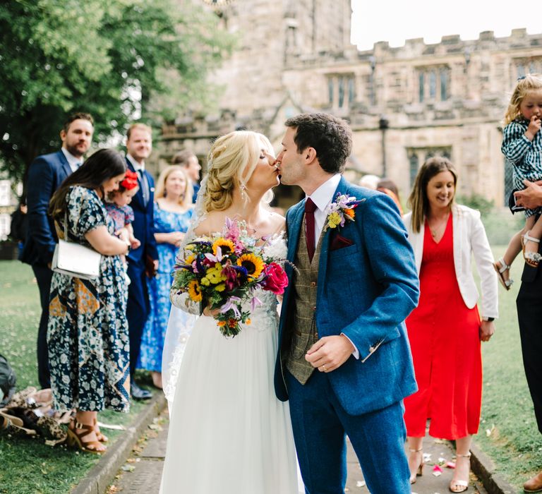 Bride holds bright floral bouquet and kisses her groom who wears matching floral buttonhole 