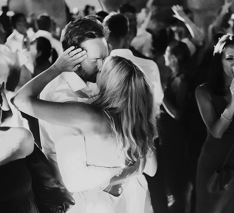 Bride and groom share a kiss ring their wedding party as guests dance 