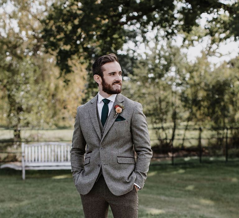 Groom in a grey tweed suit with a dark green tie and pocket square with dark brown leather shoes and a pink and red flower buttonhole 