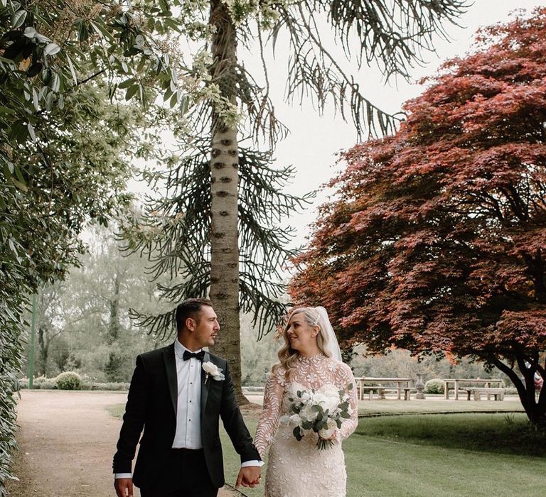 Bride in high neck lace wedding dress walks alongside her groom in black tie at Hampton Manor