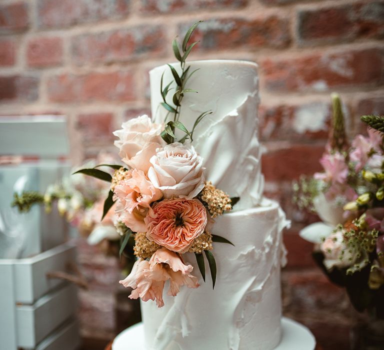 Buttercream two tiered wedding cake in off-white colour with icing details and light pink rose, foliage and golden decorations 