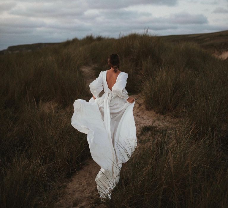 Bride walks through fields in silk floaty wedding dress with low back and bow complete with wrap styling