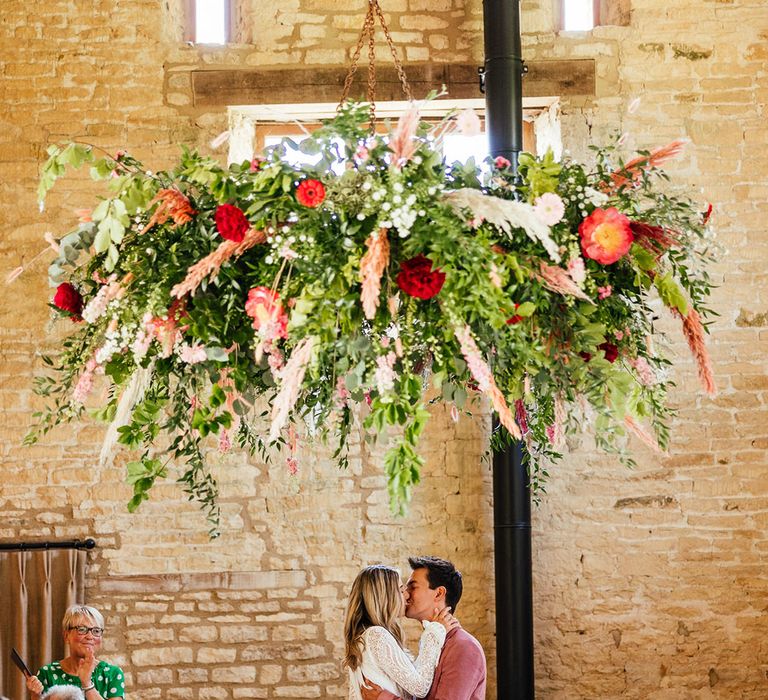 Bride in long sleeve lace boho wedding dress with tassels and groom in pink corduroy suit with pink and red rose, foliage and dried flower boutonniere kissing underneath large suspended foliage decor